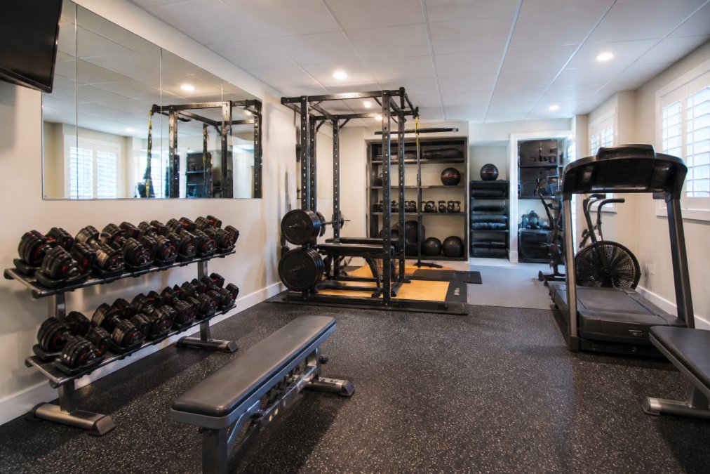 Well-organized basement home gym with multiple workout zones, squat rack in background, adjustable dumbbells displayed on wall-mounted rack, cardio equipment near ventilation, wall mirrors reflecting the space, organized storage shelves