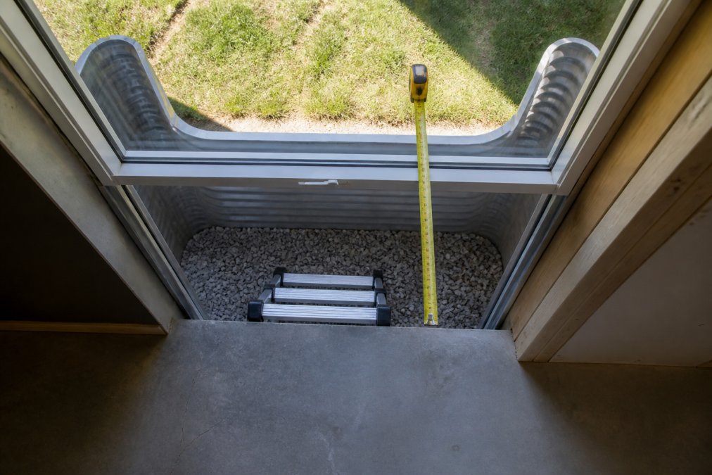 Overhead view of installed basement egress window well showing depth measurement, interior ladder rungs, drainage gravel, and the clear opening to grade with bright daylight visible