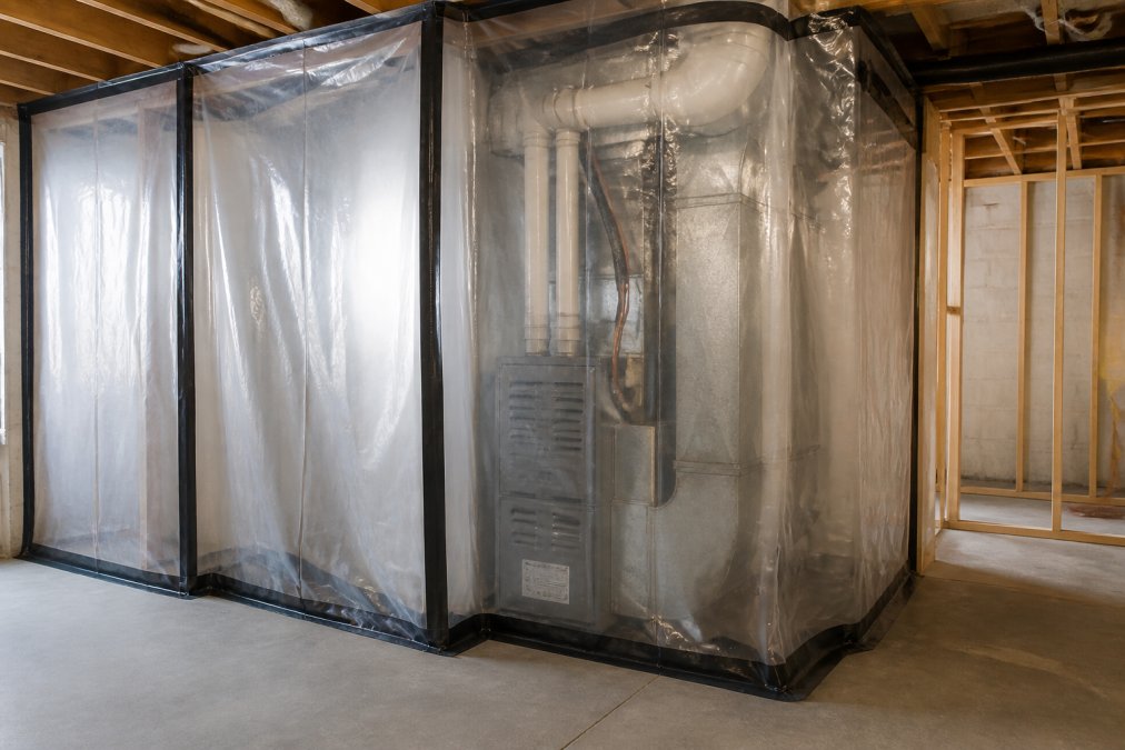 Construction site with heavy plastic sheeting sealed around furnace and return air ductwork with duct tape, plastic barriers protecting HVAC equipment from drywall dust and debris, professional containment setup