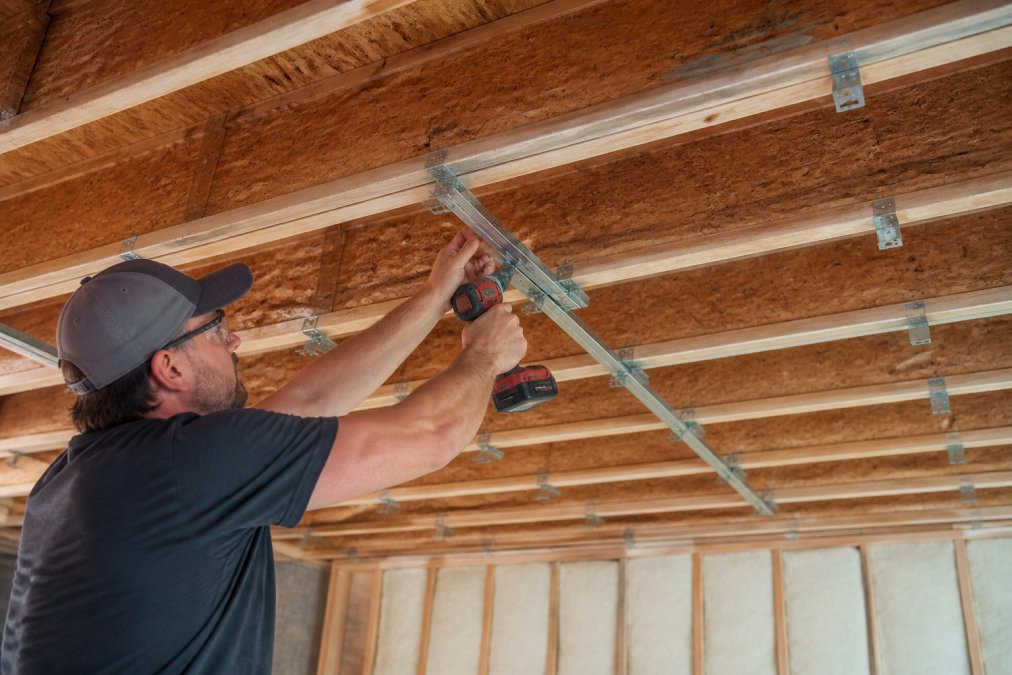Professional contractor installing resilient channels on basement ceiling framing, showing metal clips and channels mounted perpendicular to joists with proper spacing, natural workshop lighting
