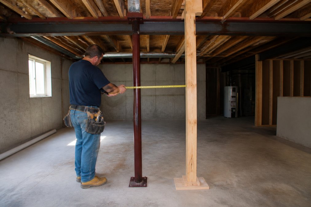 A contractor in work boots and a tool belt using a tape measure to check the spacing of a steel support column in a residential basement, exposed floor joists overhead and a temporary wood shoring post visible nearby