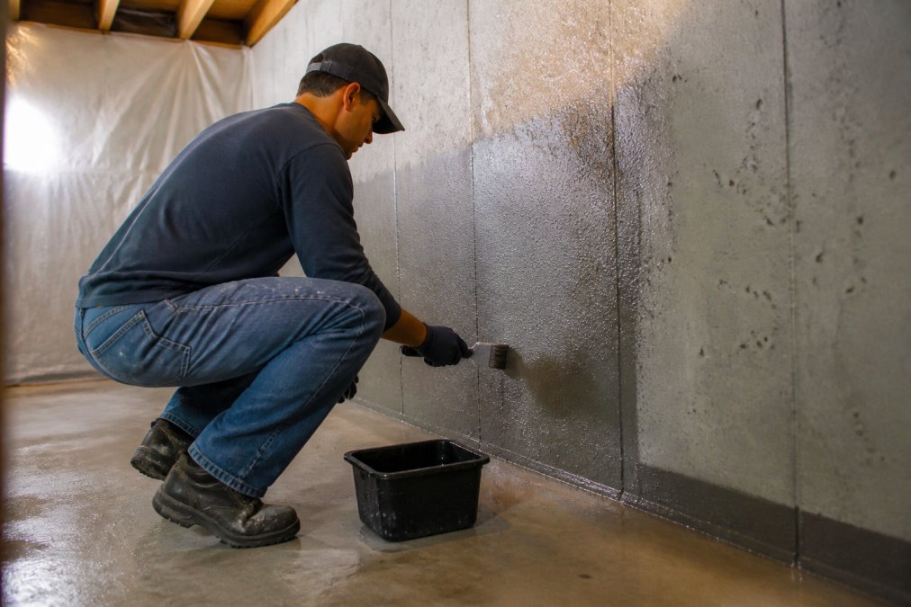 Contractor applying waterproofing sealant to basement foundation walls with brush and roller, moisture visible on concrete surface, professional work environment with protective sheeting