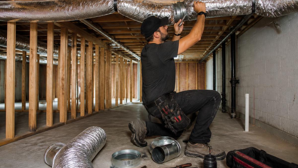 HVAC technician adjusting flexible ductwork around wooden basement joists in an unfinished basement, using a wrench to secure duct connections, with concrete floor and exposed framing visible
