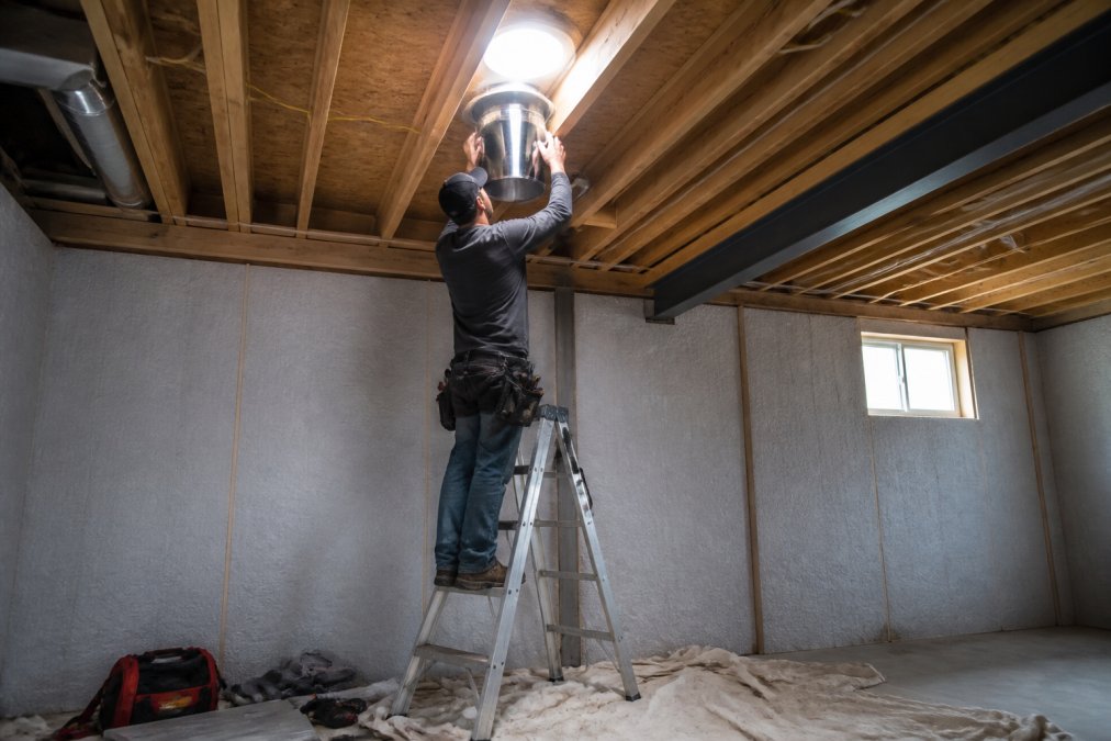 A basement contractor on a stepladder installing a solar tube in a ceiling, tools and a drop cloth below, bright daylight streaming down through the open roof flashing