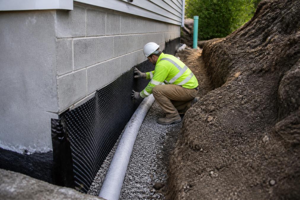 Contractor in safety gear working on exposed foundation area with visible drainage tile installation and grading work around home's perimeter in daylight