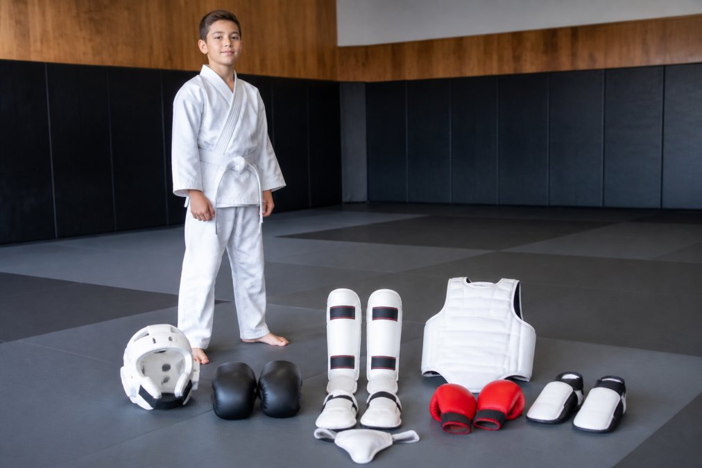 A young martial arts student in a crisp white gi and belt standing in a dojo, surrounded by sparring gear including headgear, gloves, and shin guards laid out on a mat