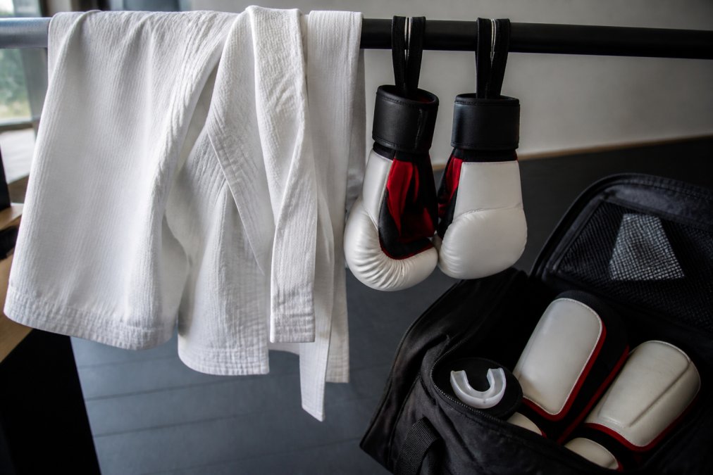 Close-up of a martial arts uniform and sparring gloves hanging to air dry on a rack, with a mouthguard and shin guards visible in an open gear bag nearby