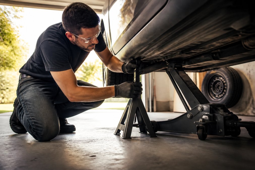 A person in a home garage placing a jack stand under the frame of a car raised on a floor jack, safety glasses on, spare tire leaning against the wall, warm afternoon light through the open garage door