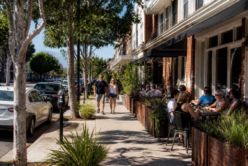 A tree-lined residential street in Burbank Rancho with local boutique shops, outdoor café seating, pedestrians strolling, and parked cars along the street on a sunny afternoon