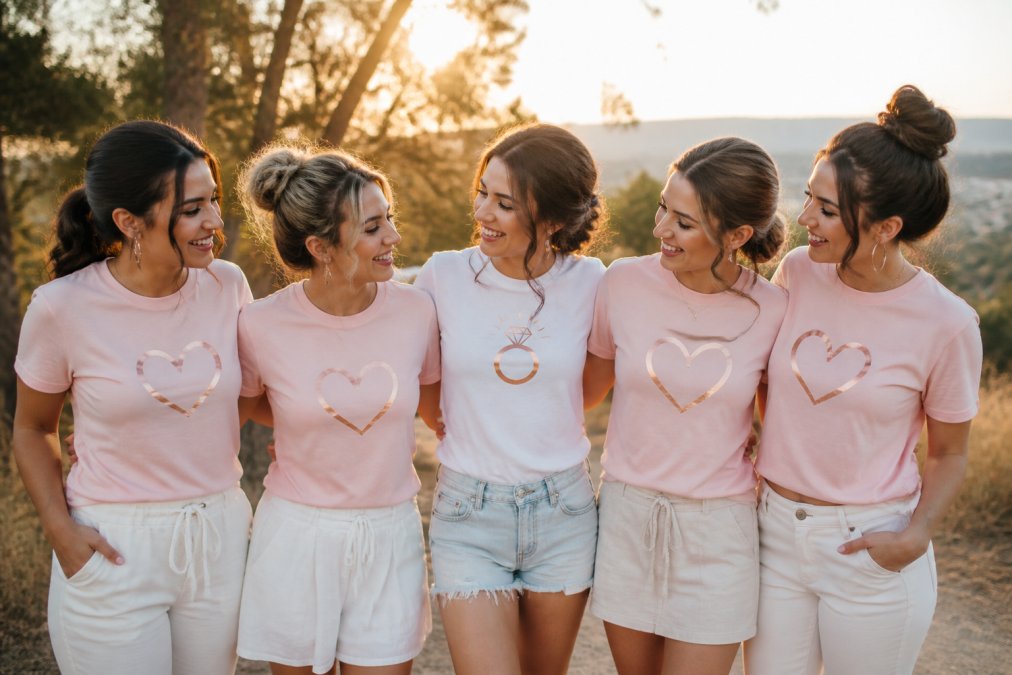 A bridal party of five women wearing coordinated custom shirts with rose gold metallic lettering and minimalist designs, posed outdoors in golden hour sunlight with soft-focus background