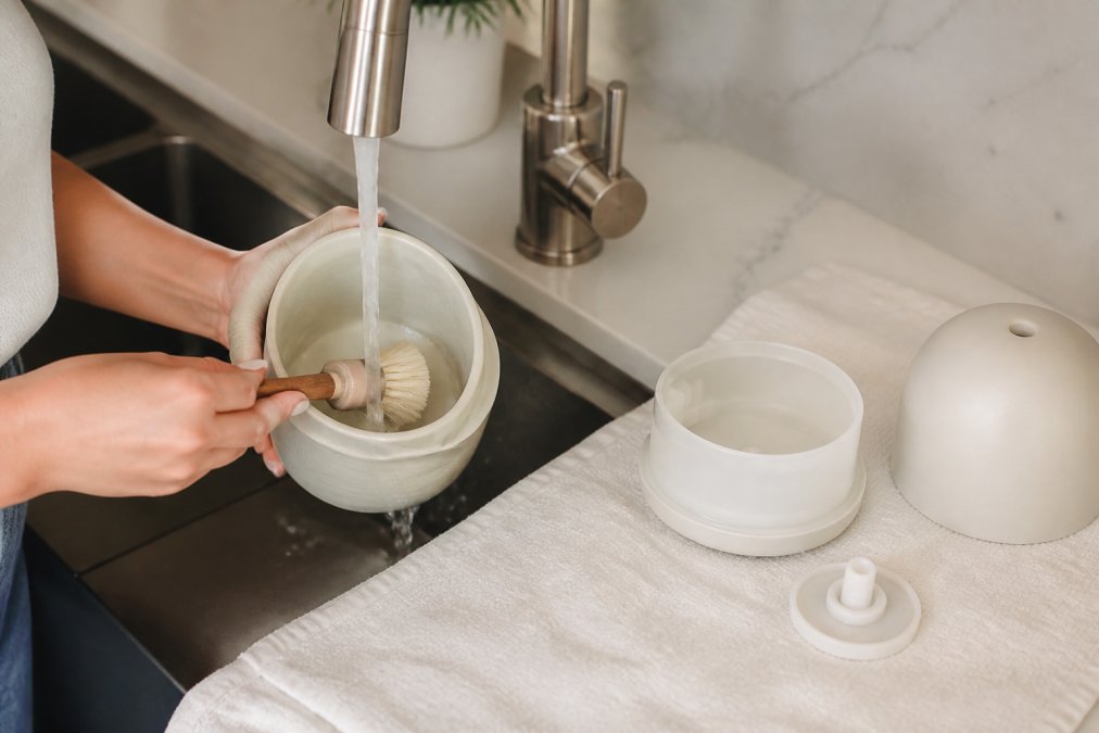 Close-up of hands gently cleaning a ceramic diffuser tank with a soft-bristled brush under running water, diffuser components laid out on a white cotton cloth, clean kitchen counter in background