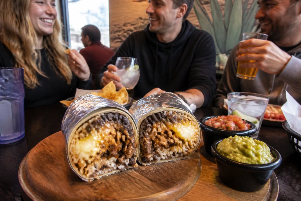 An overhead close-up of a large, foil-wrapped loaded burrito cut in half on a wooden board, revealing layers of seasoned carnitas, yellow rice, black beans, and melted cheese, with small bowls of red salsa and guacamole beside it in a vibrant, casual restaurant setting