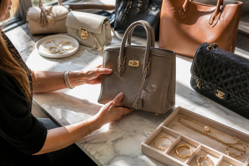 Woman's hands examining luxury handbags and delicate jewelry pieces displayed on a marble table with natural afternoon light streaming in