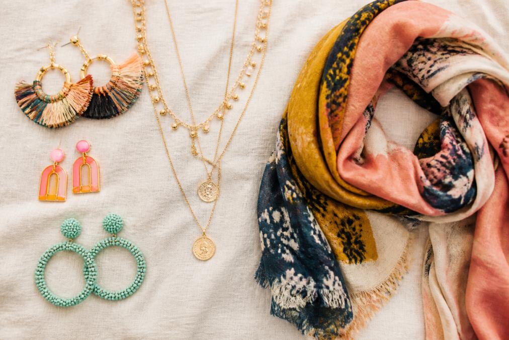 Flat lay of colorful statement earrings, layered necklaces, and patterned scarves arranged on a soft neutral linen background