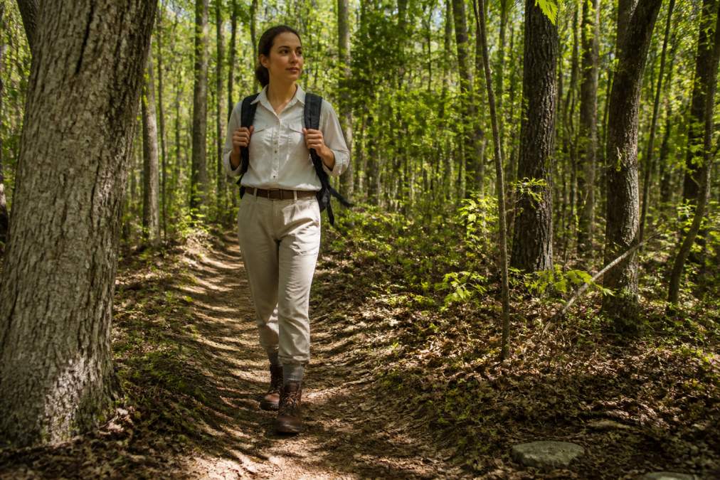 A hiker in light-colored long pants tucked into socks and a long-sleeved shirt walking a narrow forest trail, mid-step, with dappled sunlight through the trees