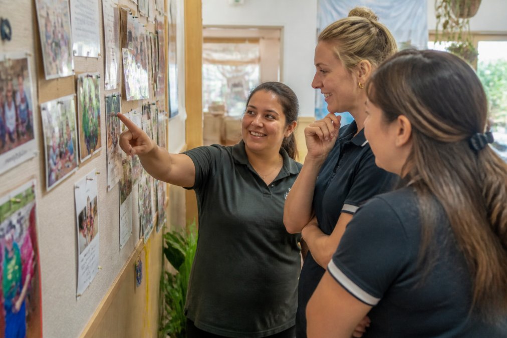 Early childhood educators gathered around a notice board displaying child safety values and recent examples of protective behaviours in action, with educators pointing and discussing the posted content