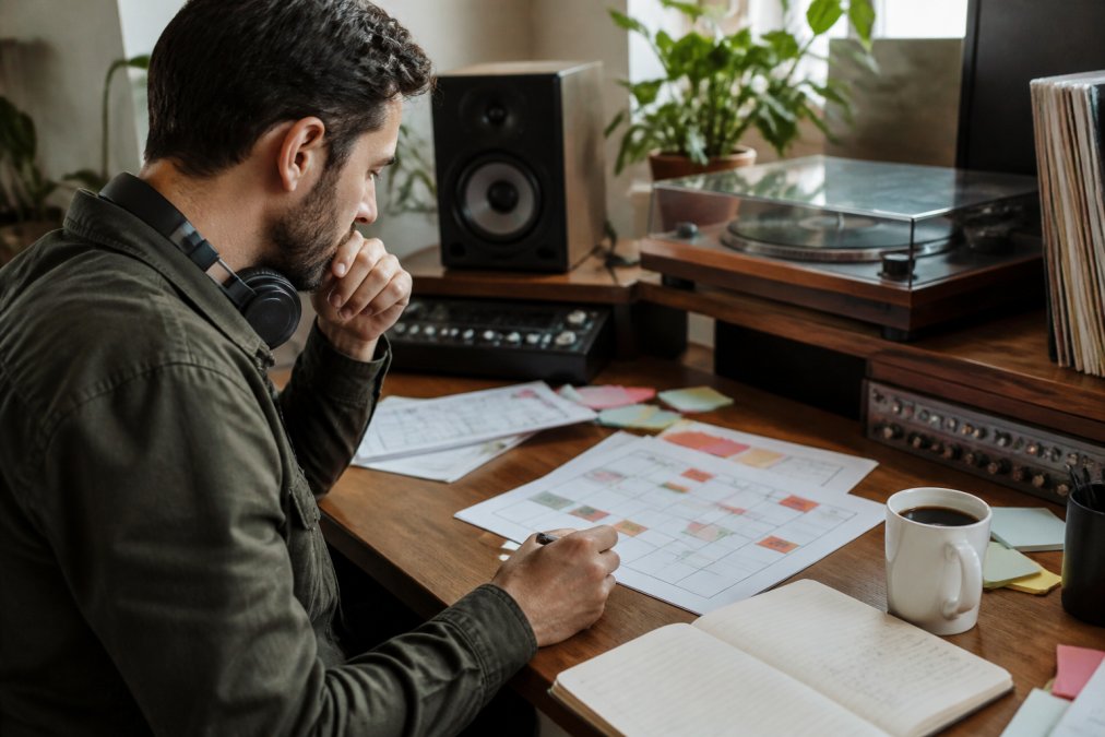 Music producer reviewing a detailed release calendar spreadsheet on a desktop monitor with studio headphones and production notes scattered on the desk beside a coffee cup