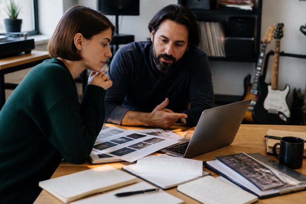 Record label manager and artist collaborating over a laptop in a modern office, reviewing a detailed release strategy document with color-coded timelines, marketing assets displayed on the screen, and notebooks with handwritten notes scattered nearby