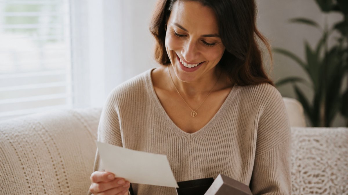 Woman wearing an elegant personalized necklace with delicate chain, holding a custom message card in soft natural light, smiling with genuine emotion as she reads the personal message
