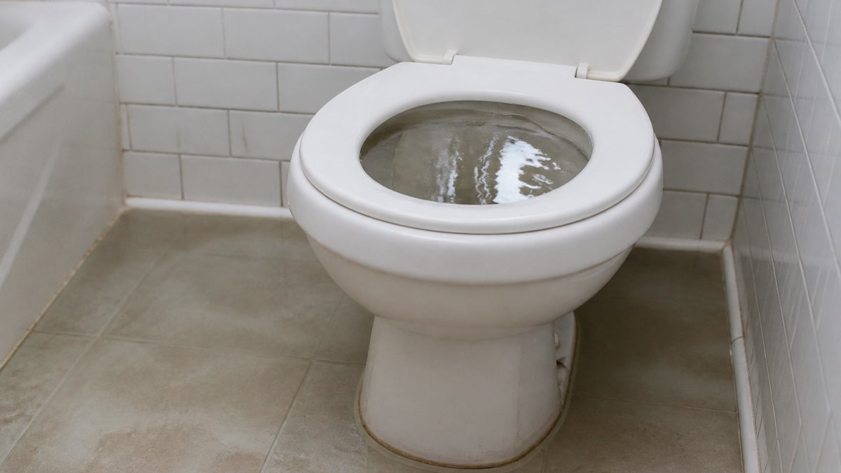 Backed-up toilet bowl with water pooling from signs of broken sewer pipes in a residential bathroom