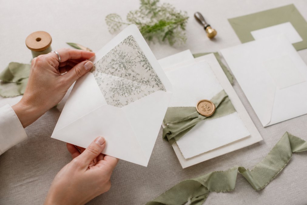 Hands lifting an invitation envelope flap to reveal a patterned liner with a wax-sealed suite on linen