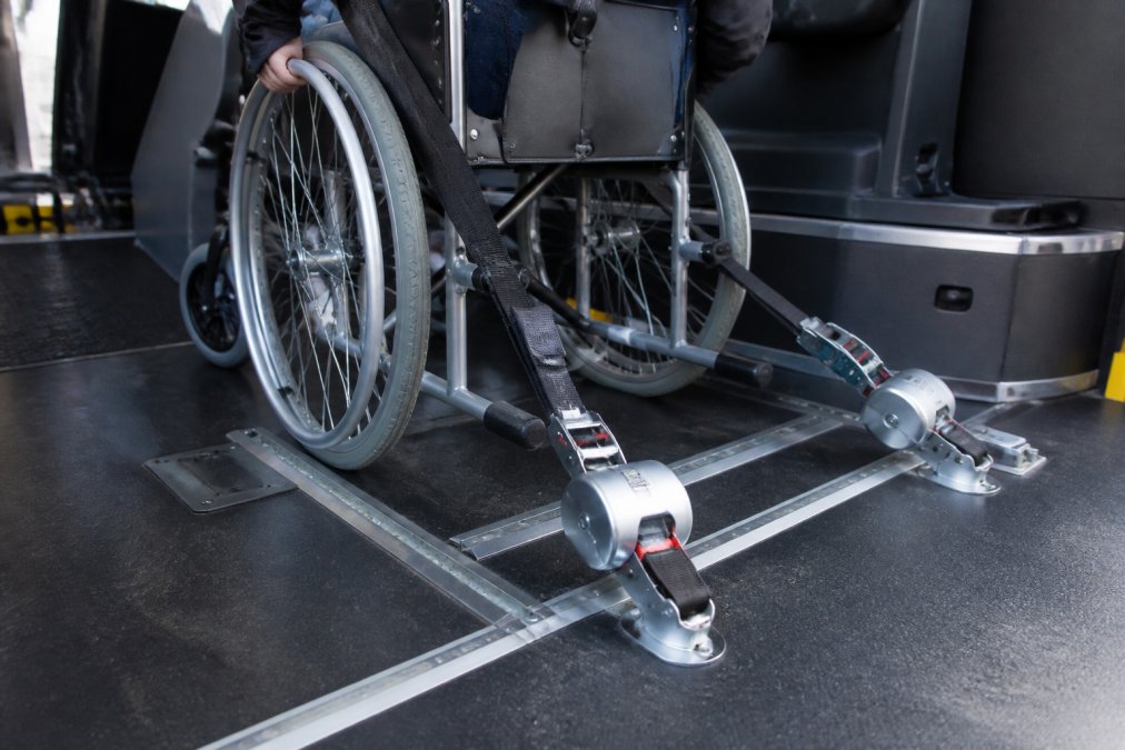 Close-up van een rolstoel die veilig is vastgezet in een aangepaste taxi met zichtbare beveiligingssystemen, stalen beugels en driepuntsgordels die de rolstoel op zijn plaats houden