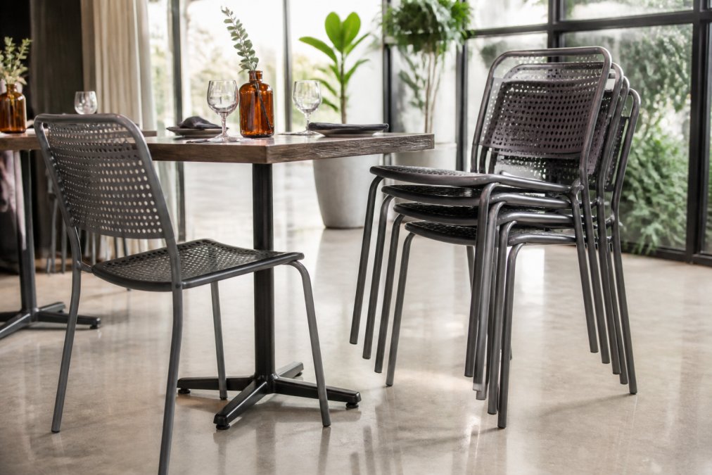 A neat stack of grey perforated metal chairs beside a wooden bistro table in a bright modern cafe, natural daylight from large windows, polished concrete floor