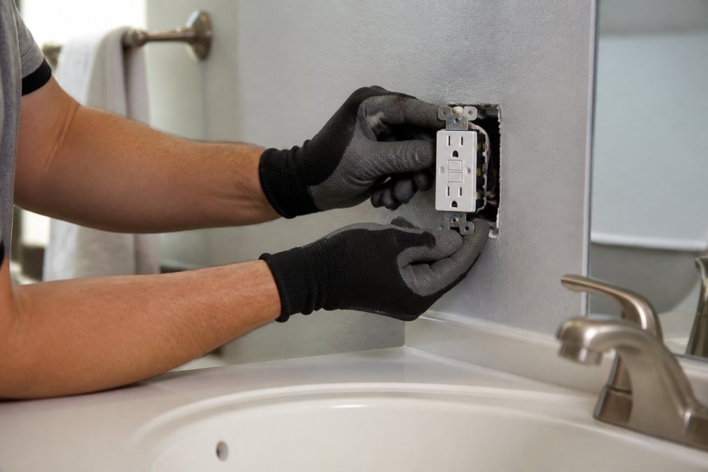 Close-up of a licensed electrician's gloved hands installing a white GFCI outlet with visible test and reset buttons into a bathroom wall near a porcelain sink, under bright overhead lighting