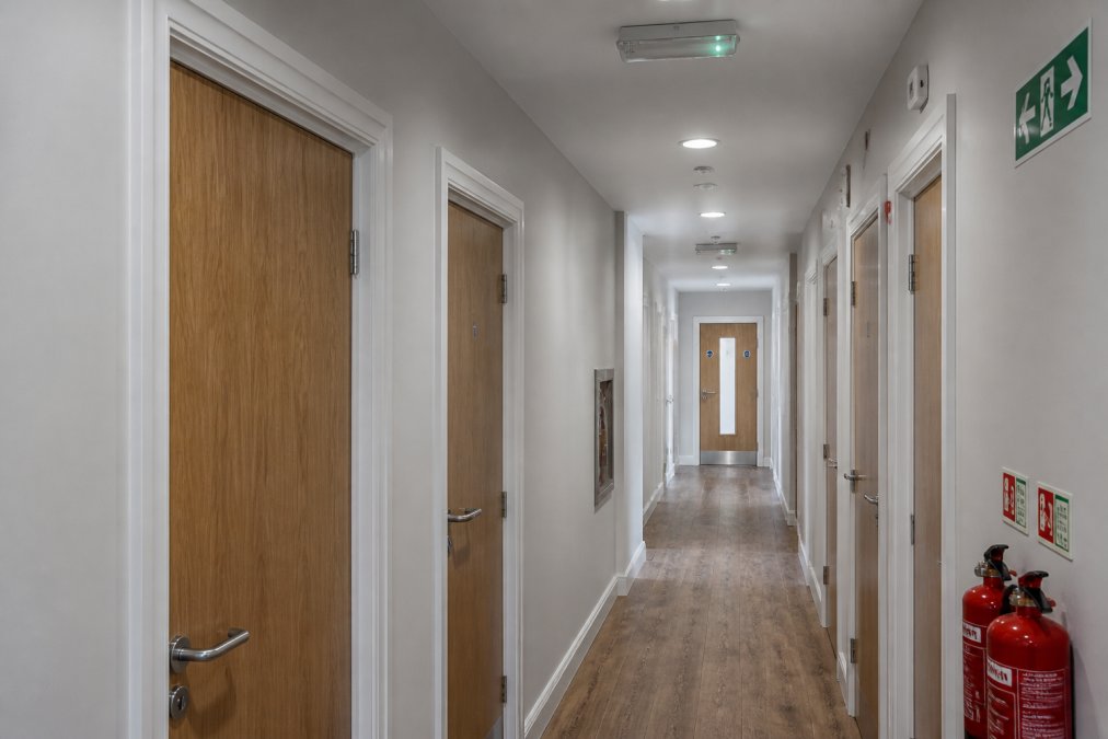 Interior corridor of a modern HMO conversion showing multiple bedroom doors with fire-resistant frames, adequate emergency lighting, clear escape route signage, and visible fire safety equipment on neutral-coloured walls