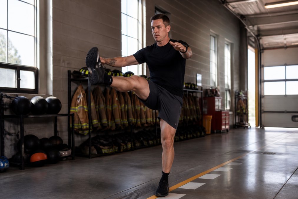 Firefighter in workout gear performing dynamic leg swings and mobility drills in a firehouse training bay with natural light streaming through tall windows
