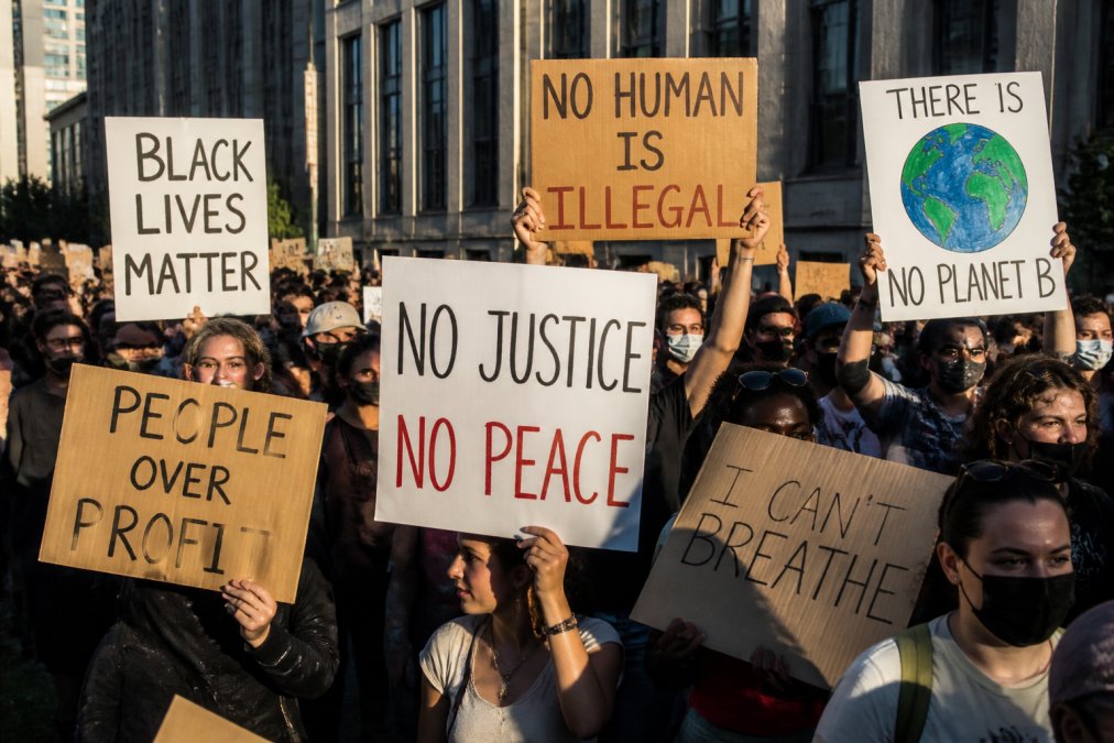 A diverse crowd at a modern protest holding up a variety of creative cardboard signs with bold slogans, seen from a slight low angle, late afternoon sunlight casting long shadows