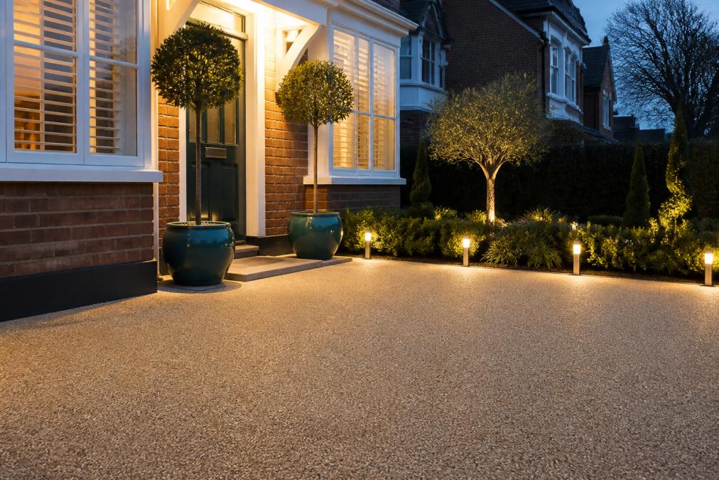 A neat front garden of a British home with a smooth resin bound driveway, evergreen shrubs in glazed pots, and solar path lights glowing at dusk