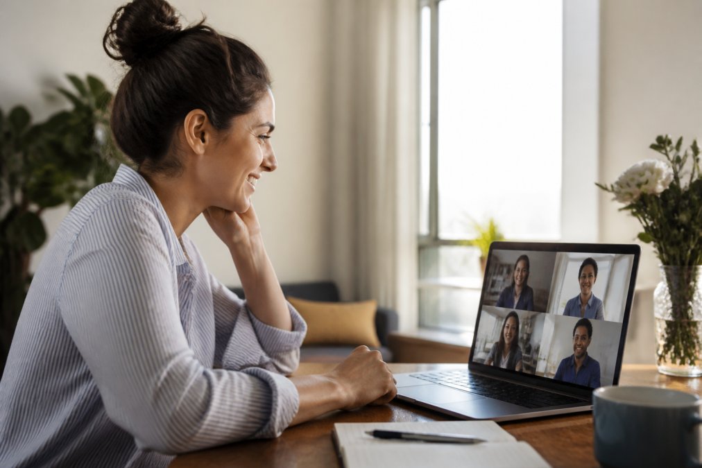 Remote employee smiling at laptop during a virtual team video call, with colleagues visible on screen in the background, warm office lighting streaming through a window