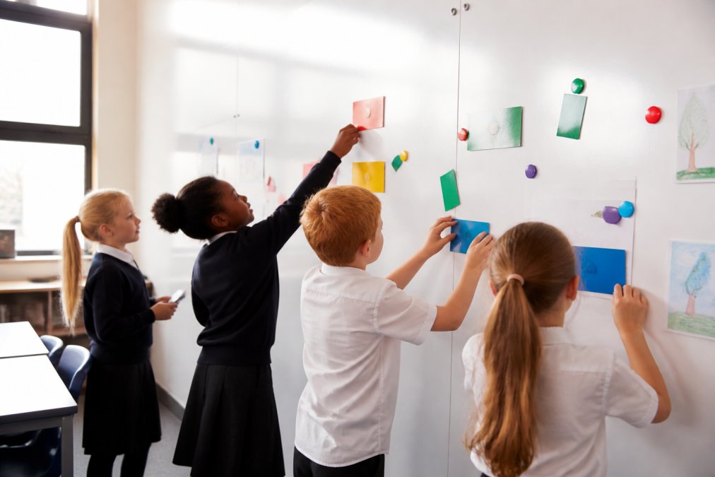 Primary school pupils collaborating at a magnetic whiteboard wall, attaching colourful cards and writing notes during a group activity in a bright classroom