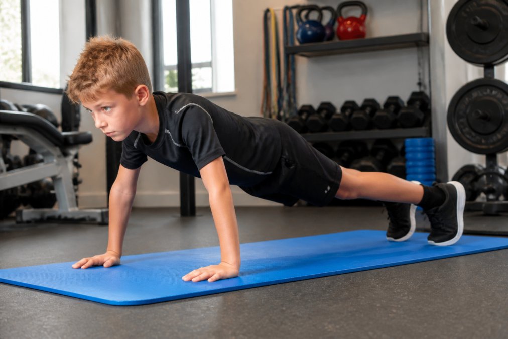 Young child performing a plank hold on a blue exercise mat in a bright home gym, maintaining straight body alignment with hands shoulder-width apart and core engaged, demonstrating proper form
