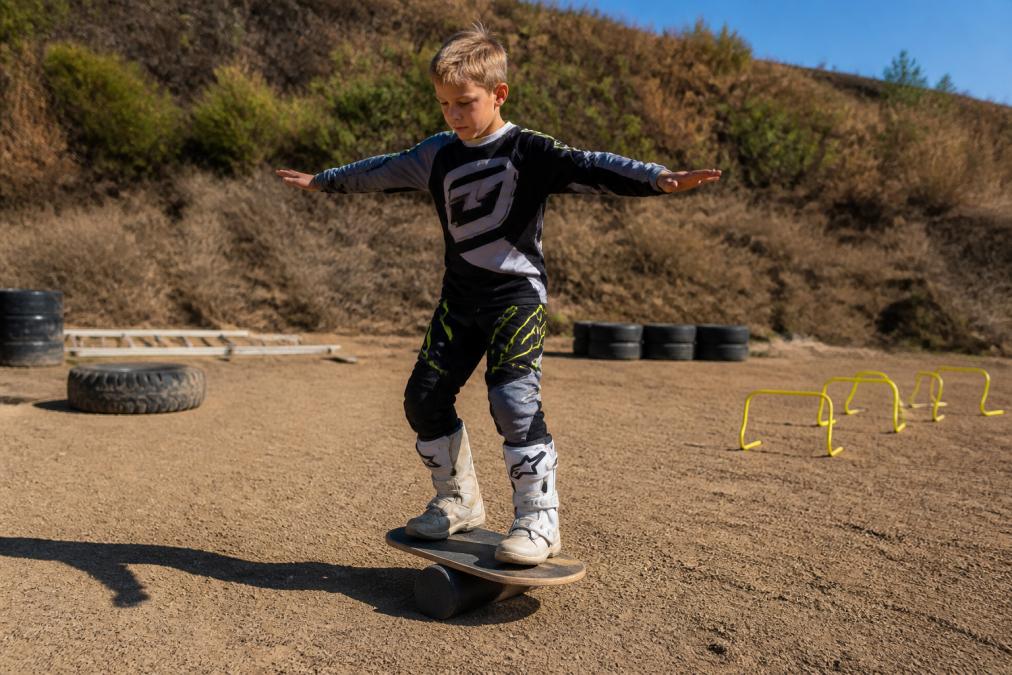 Child standing on a balance board with arms outstretched for stability, concentrating intently while maintaining equilibrium on the unstable surface in a bright training area