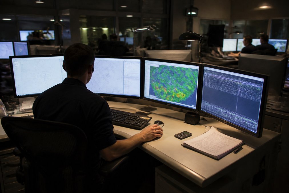 Aircraft dispatcher seated at a curved desk with four monitors showing flight plans, weather radar, and radio logs in a dim airline operations center, colleagues working at nearby stations