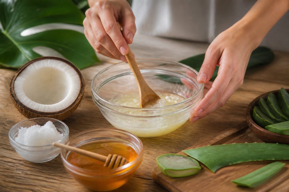 Hands mixing natural ingredients in a glass bowl: coconut oil, honey, and aloe vera, with fresh ingredients arranged on a wooden surface