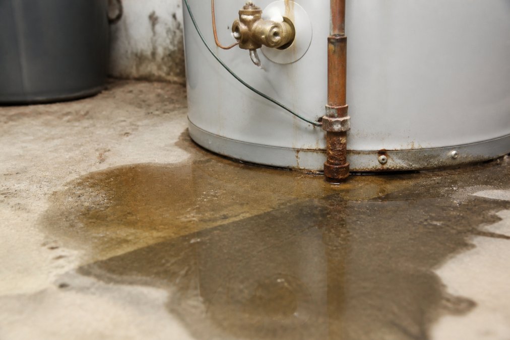 Close-up of water pooling around the base of a residential water heater tank, showing moisture and corrosion on metal connections