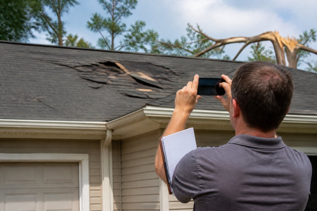 Homeowner photographing roof damage with smartphone from ground level, showing visible storm damage like missing shingles and dents, with notepad and pen nearby for documenting details