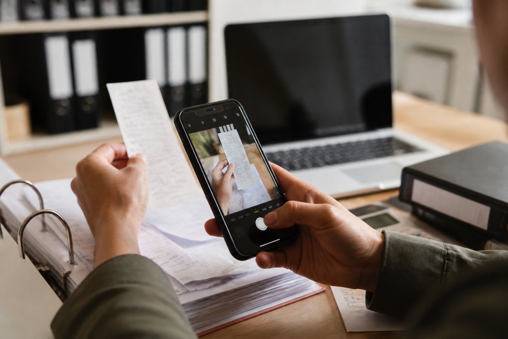 Nahaufnahme von Händen, die Belege mit einem Smartphone scannen, auf einem Schreibtisch mit Ordnern und Dokumenten, digitale Steuererklärung auf dem Bildschirm