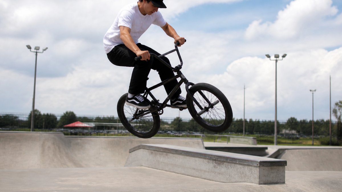 BMX rider wearing a snapback cap mid-trick at an outdoor skatepark, hat positioned securely during aerial maneuver with curved bill visible, natural sunlight casting shadows on concrete