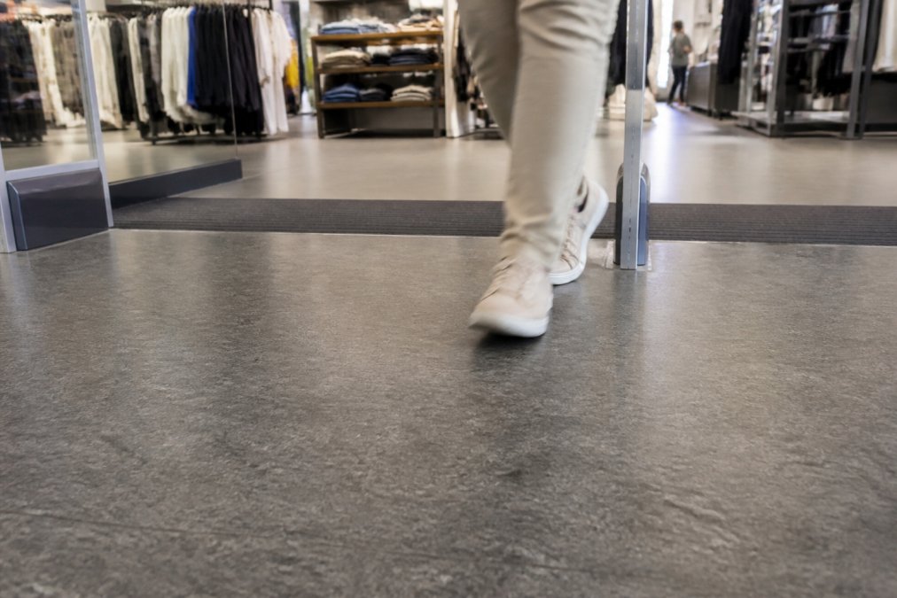 10 ting du skal vide om PVC gulve 2 Close-up of a durable PVC floor at a busy retail store entrance, surface texture and wear layer visible under bright ceiling lighting, a customer walking past in the background