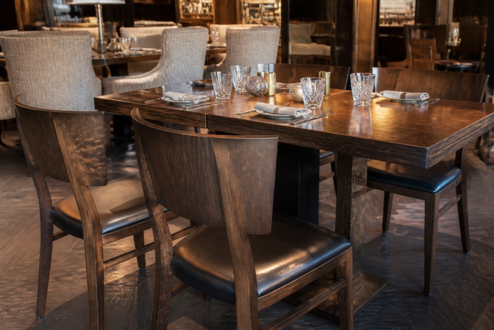 Elegantes Restaurant-Interieur mit massiven Massivholzstühlen und einem Tisch aus dunklem Holz, der in warmem Licht erstrahlt, im Hintergrund weitere Tische mit edlen Stoffbezügen