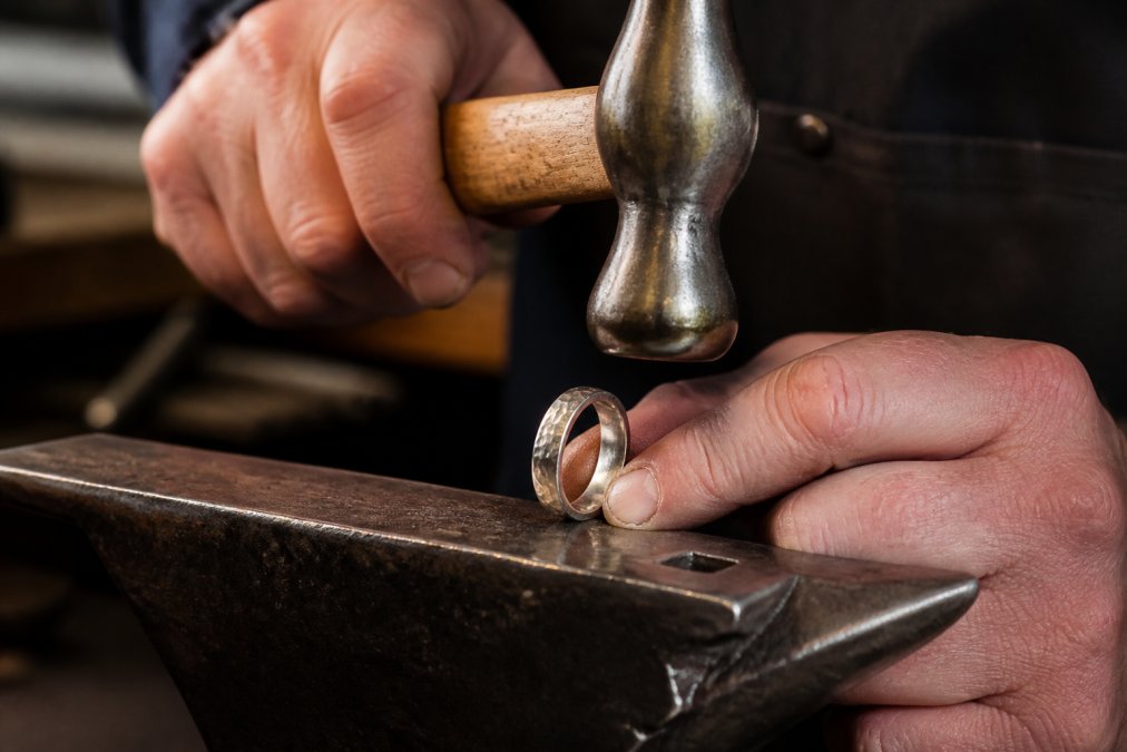 Close-up of a master jeweller's hands carefully hammering texture into a white gold wedding band on an anvil, with soft workshop lighting illuminating the metalwork and fine detail