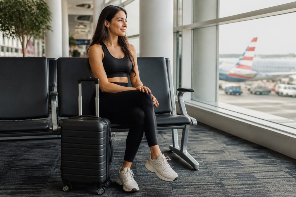 A woman in a black seamless activewear set sitting in a bright airport terminal, a small carry-on suitcase at her feet, looking relaxed and ready to board