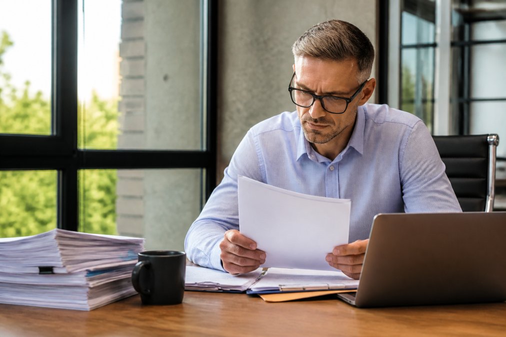 Real estate investor reviewing a stack of loan documents and financial statements at a modern desk, laptop open to a spreadsheet, warm afternoon light through office windows