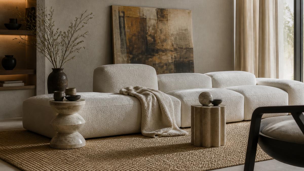 A softly lit modern living room with a low-profile modular sofa in cream boucle, styled with a textured throw, two sculptural side tables and a large piece of art leaning against the wall