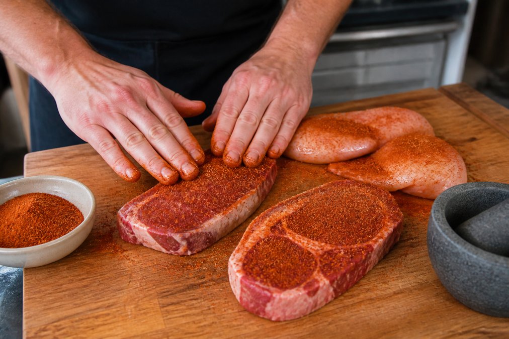 Close-up of raw steaks and chicken breasts being rubbed with red chile powder spice blend on a wooden cutting board, hands applying the seasoning generously