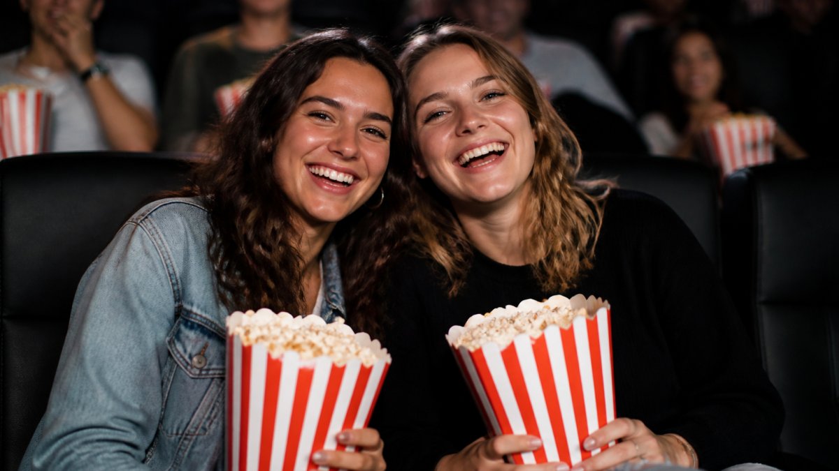 Two best friends laughing together at a movie theater, holding popcorn and smiling at the camera in a warm, friendly moment